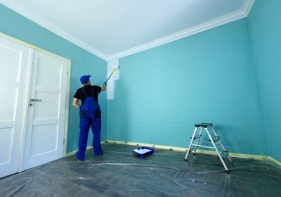 Man in a working overall is painting the wall in white color